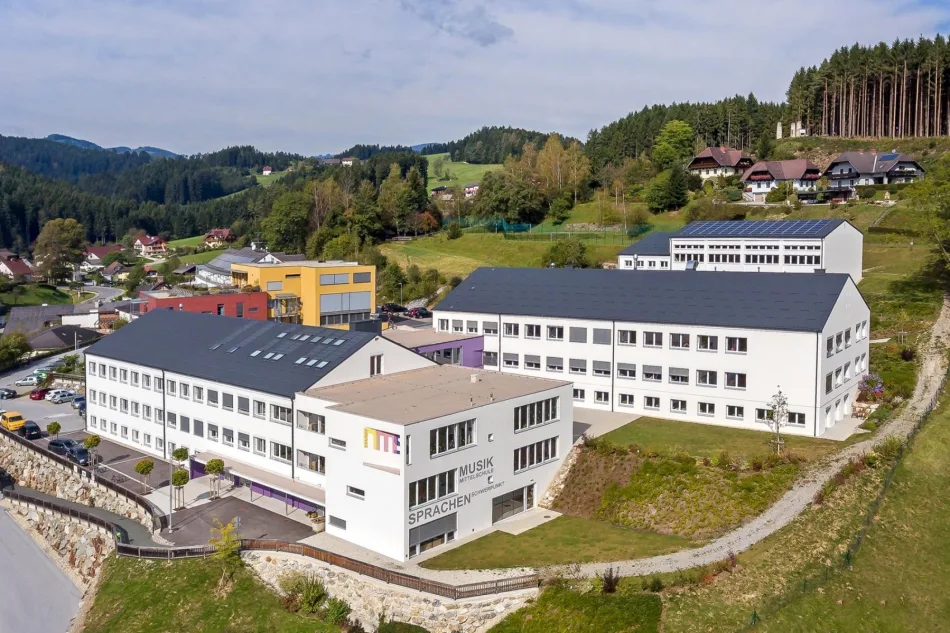 Foto: Mittelschule Birkfeld Luftbild der Mittelschule Birkfeld: weißes Schulgebäude am Hang, dahinter Wiesen und Wälder des Feistritztals