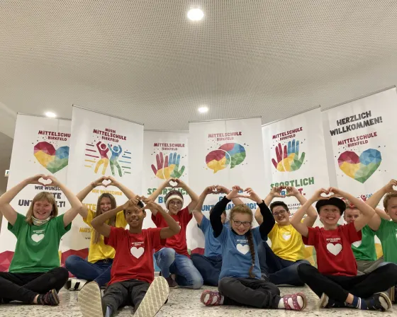 Schülerinnen und Schüler in roten, gelben, grünen und blauen T-Shirts formen mit den Händen Herzen vor den „Wachse BUNT.“-Fahnen