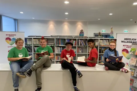 Fünf Kinder in bunten T-Shirts sitzen mit Büchern in der Schulbibliothek